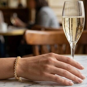 Puede incluir: Una pulsera dorada con piedras preciosas redondas y transparentes adorna una mu&ntilde;eca. Una copa de champ&aacute;n se encuentra sobre una mesa de m&aacute;rmol. El fondo est&aacute; borroso, sugiriendo un restaurante o bar.