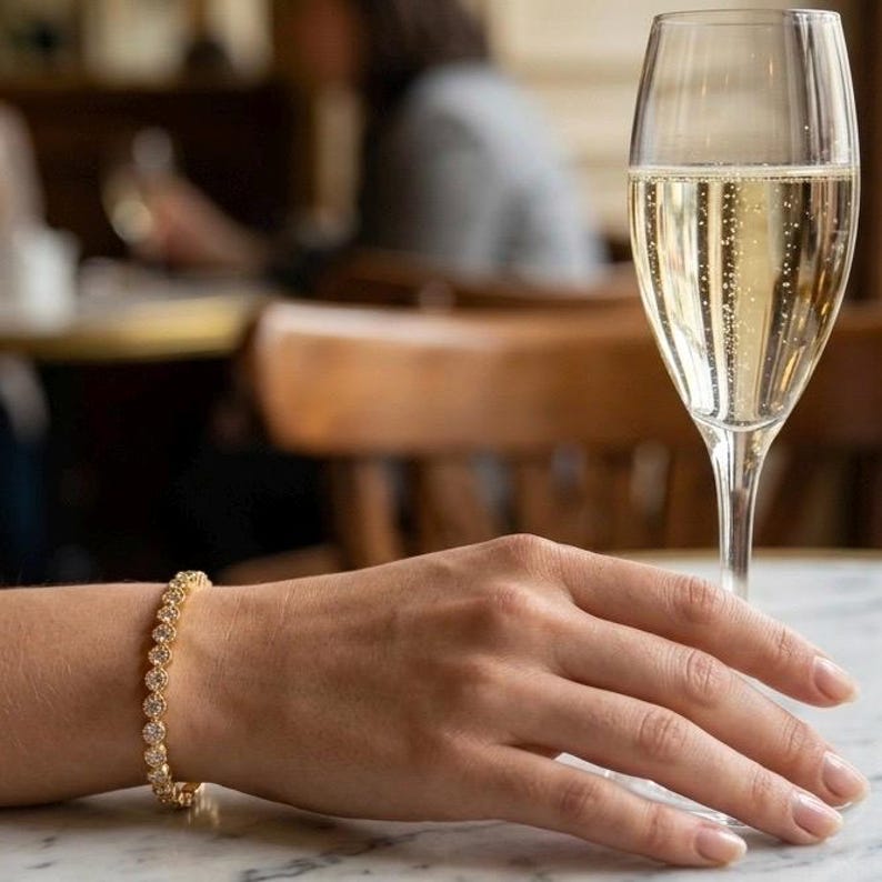 Puede incluir: Una pulsera dorada con piedras preciosas redondas y transparentes adorna una mu&ntilde;eca. Una copa de champ&aacute;n se encuentra sobre una mesa de m&aacute;rmol. El fondo est&aacute; borroso, sugiriendo un restaurante o bar.
