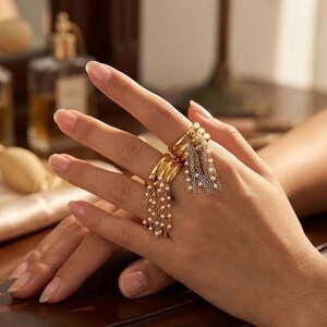May include: Two gold rings with dangling chains of small pearls and silver chains. The rings are worn on a hand, showcasing the jewelry's design and texture. The rings have a layered design.