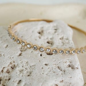 Puede incluir: Una pulsera dorada con una fila de cristales claros y brillantes. La pulsera descansa sobre una superficie texturizada de color blanquecino, creando un contraste entre el metal liso y el fondo rugoso.