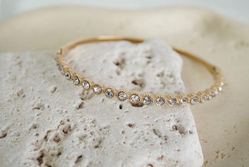 Puede incluir: Una pulsera dorada con una fila de cristales claros y brillantes. La pulsera descansa sobre una superficie texturizada de color blanquecino, creando un contraste entre el metal liso y el fondo rugoso.