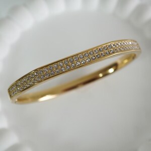 May include: A gold-colored bangle bracelet with two rows of small, clear crystals. The bracelet has a slightly curved design and a polished finish, reflecting light. The crystals are evenly spaced, adding sparkle to the jewelry.