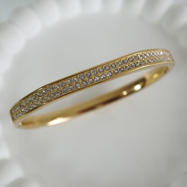 May include: A gold-colored bangle bracelet with two rows of small, clear crystals. The bracelet has a slightly curved design and a polished finish, reflecting light. The crystals are evenly spaced, adding sparkle to the jewelry.