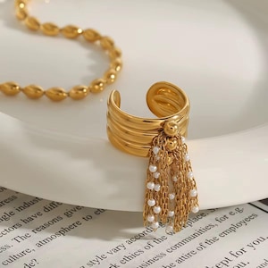 May include: A gold-colored ring with a layered band design and a dangling tassel of small white beads and gold chains. The ring is open-ended and sits on a white surface. A gold necklace is visible in the background.