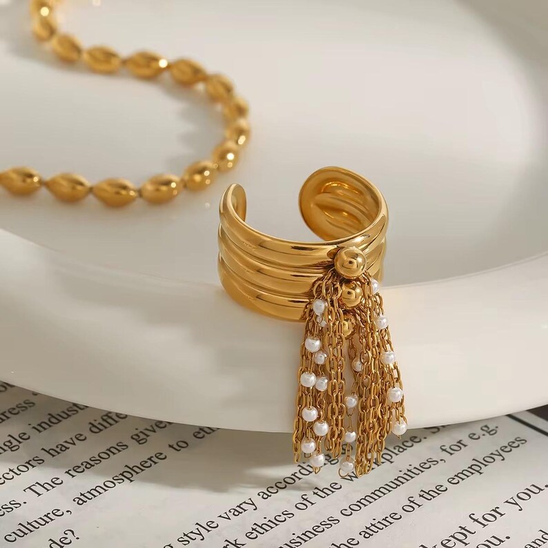 May include: A gold-colored ring with a layered band design and a dangling tassel of small white beads and gold chains. The ring is open-ended and sits on a white surface. A gold necklace is visible in the background.