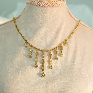 May include: A gold-colored necklace with multiple dangling clear crystal pendants. The necklace is displayed on a pink mannequin, showcasing its delicate design and sparkling gemstones. The necklace has a delicate chain.