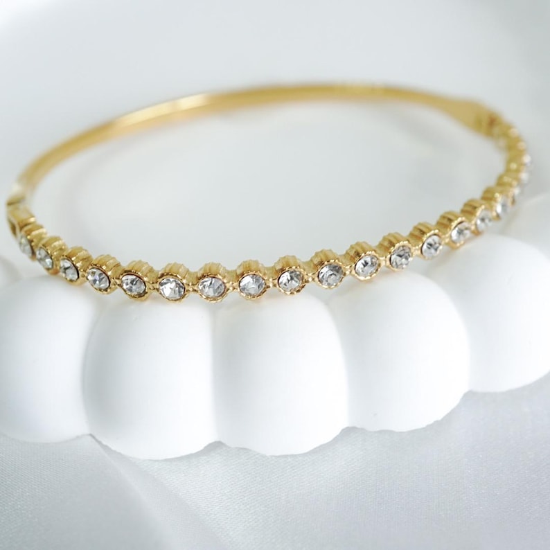 Puede incluir: Una pulsera dorada con una fila de piedras preciosas redondas y transparentes. La pulsera descansa sobre una superficie blanca en forma de nube, con un fondo de tela blanca.