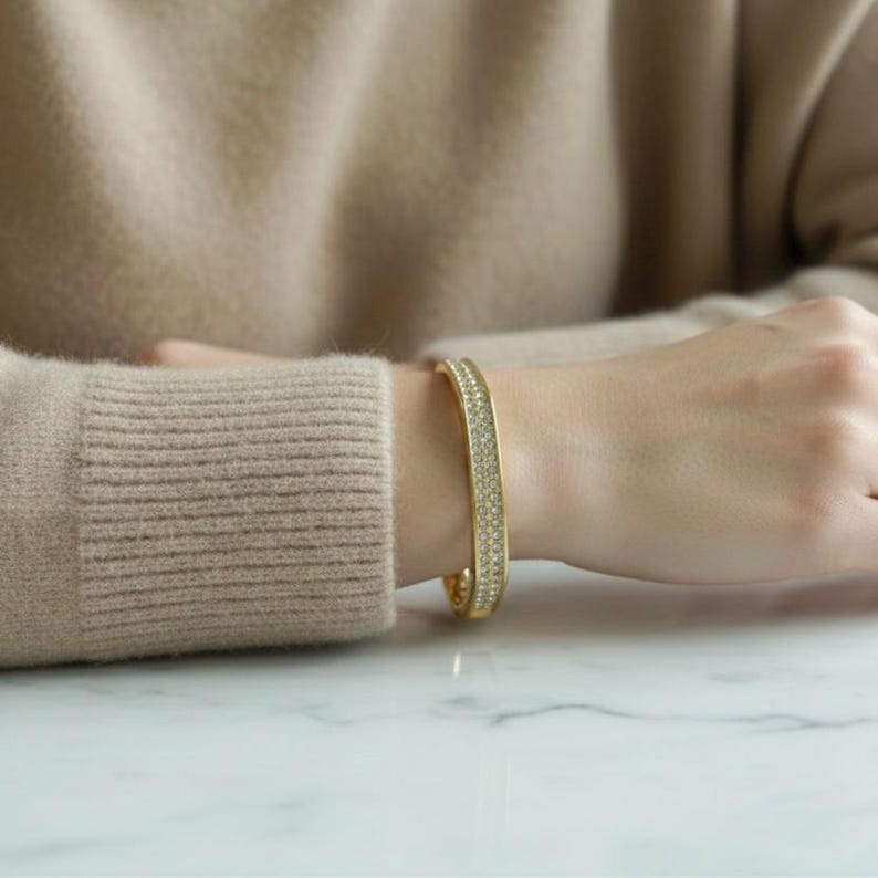 May include: A gold-colored bracelet with a row of small, sparkling crystals. The bracelet is worn on a wrist, with a neutral-toned sweater in the background. The bracelet appears to be approximately 2.5 inches in diameter.