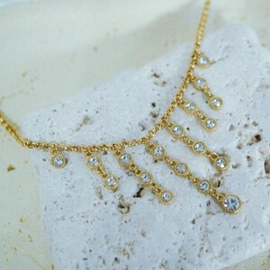 May include: A gold-colored necklace with dangling, clear crystal accents. The necklace features a delicate chain and multiple crystal pendants, creating a sparkling, elegant design. The crystals are round and set in a decorative bezel.