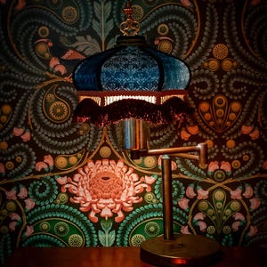 May include: A vintage-style table lamp with a blue velvet shade and a decorative gold finial. The lamp has a bronze-colored adjustable arm and base. The background features a dark floral wallpaper with shades of green, pink, and gold.