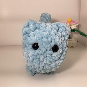 May include: A light blue, handmade crochet cat toy with large black eyes. The plush toy has a textured, fluffy appearance and a small tail. The cat toy is sitting on a white surface.