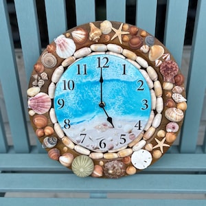 May include: A round wall clock with a beach theme. The clock face features a blue and white ocean design. The clock is surrounded by various seashells, starfish, and small stones, creating a coastal aesthetic.