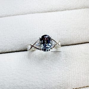 Op de afbeelding: Zilveren ring met een ovale, donkerblauwe edelsteen met paarse ondertonen. De ring heeft een gedraaid bandontwerp, met twee kleinere, driehoekige, heldere edelstenen aan weerszijden van de hoofdsteen. Op een witte achtergrond.