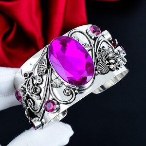May include: A silver cuff bracelet featuring a large, oval-shaped, faceted magenta gemstone in the center. The metal surface is decorated with intricate, raised floral and scrollwork patterns, accented by several smaller, round magenta gemstones.