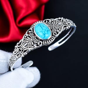 May include: Silver cuff bracelet with an oval turquoise stone. The bracelet has an open design with detailed floral and vine patterns. The turquoise stone is a bright blue, and the metal has a polished finish. The bracelet is approximately 17 cm in circumference.