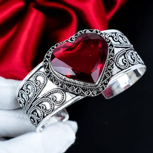 Può includere: Un bracciale a fascia in argento con una grande pietra preziosa rosso scuro sfaccettata a forma di cuore al centro. La fascia metallica è decorata con intricati motivi in filigrana che si estendono lungo i lati del bracciale.