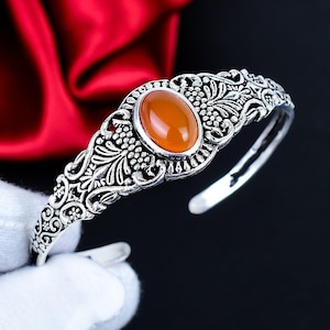 Puede incluir: Brazalete de plata adornado con una piedra preciosa ovalada de color naranja. El brazalete tiene un diseño floral detallado con una estética vintage. El diseño abierto facilita su uso. Una joya llamativa.