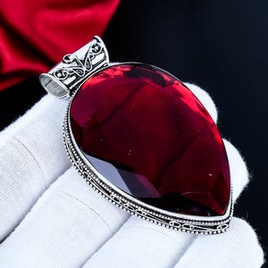 Può includere: Un grande pendente a goccia con una gemma rossa scura sfaccettata. La gemma è incastonata in una montatura in metallo color argento con dettagli intricati. Il pendente è mostrato su uno sfondo bianco.