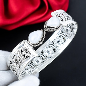 Peut inclure: Un bracelet manchette argenté avec des motifs floraux et des volutes. Il comporte trois pierres précieuses blanches en forme de poire. Les détails complexes et la finition brillante créent un bijou saisissant.