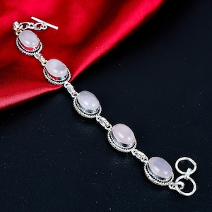 Könnte beinhalten: Ein Silberarmband mit fünf ovalen, blassrosa Rosenquarz-Edelsteinen. Jeder Stein ist in einen gedrehten Silberrahmen gefasst und durch Silberglieder verbunden. Das Armband verfügt über einen Knebelverschluss mit einem T-Stab und einem Ring.