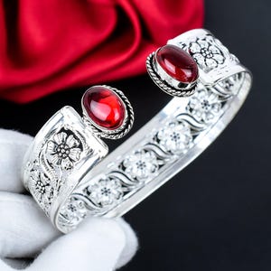 Può includere: Bracciale a polsino in argento con due pietre preziose rosse ovali. Il bracciale presenta motivi floreali e a volute. Le pietre preziose sono incastonate in una lunetta d'argento. Il bracciale è tenuto da una mano guantata.