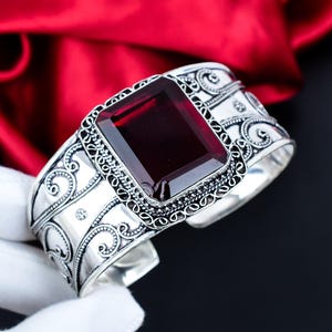 Könnte beinhalten: Ein silbernes Armreif-Armband mit einem großen, rechteckigen, tiefroten Edelstein. Das Armband hat aufwendige Schnörkel und dekorative Details um den Edelstein und am Band. Der Hintergrund ist ein reichhaltiger roter Stoff.