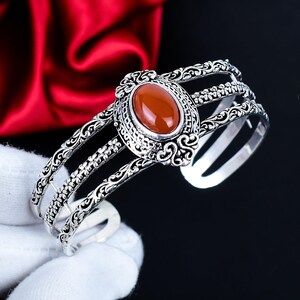 Peut inclure: Bracelet manchette argenté avec une pierre centrale ovale brun-rouge. Le bracelet présente des motifs floraux et de volutes complexes. Le bracelet manchette a trois bandes parallèles et est présenté sur fond noir.