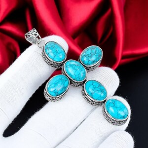 Puede incluir: Un colgante plateado en forma de cruz, con seis piedras turquesas ovaladas. Las piedras están engastadas en biseles ornamentados y detallados. El colgante está sostenido por una mano enguantada sobre un fondo de satén rojo.