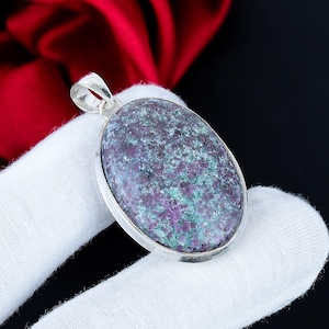 Puede incluir: Colgante ovalado con engaste plateado. El colgante presenta una piedra moteada con tonos morados, verdes y grises. La piedra está engastada en un marco plateado pulido con un lazo para una cadena.