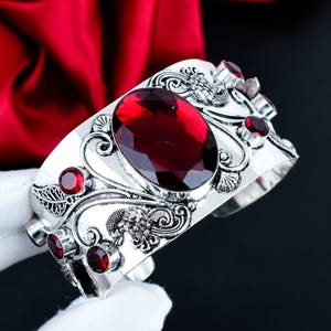 Puede incluir: Un brazalete de plata con una gran piedra preciosa de color rojo intenso y corte ovalado en el centro, rodeada de piedras rojas redondas más pequeñas. La superficie metálica presenta intrincados patrones de volutas y hojas en relieve.