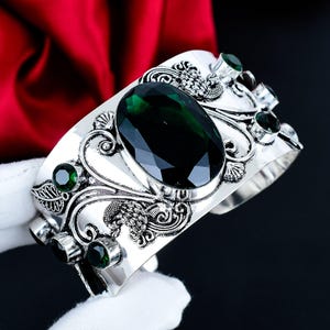 Peut inclure: Un bracelet manchette en argent orné d'une grande pierre précieuse vert foncé de taille ovale au centre, entourée de volutes métalliques complexes et de motifs de feuilles. Des pierres vert foncé rondes ornent les bords du large bracelet.
