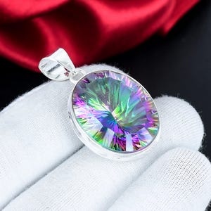 May include: A silver pendant featuring an oval-cut, iridescent gemstone. The gemstone shimmers with colours like green, purple, and blue. The pendant is held by a gloved hand, set against red fabric and a black background.