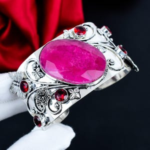 Può includere: Un bracciale a fascia in argento con una grande pietra preziosa ovale sfaccettata color rosa acceso al centro. La fascia in metallo è decorata con intricati motivi floreali e a farfalla, accentuati da diverse pietre rosse scure.