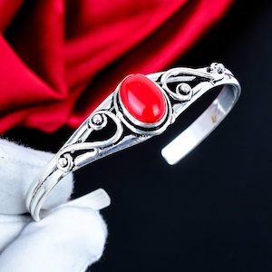 May include: A silver cuff bracelet featuring a central oval-shaped red stone. The metal band displays intricate swirling wirework patterns that extend from the stone setting towards the open ends of the bracelet, creating a decorative frame.