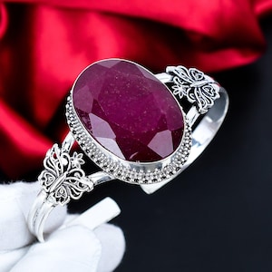 Puede incluir: Una pulsera de plata con una gran gema de rubí de corte ovalado. El rubí es de un color rojo intenso y rico y está engastado en un bisel de plata detallado. La pulsera tiene detalles decorativos de mariposas a cada lado.