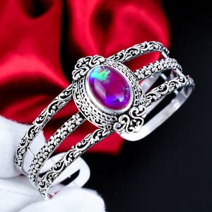 Puede incluir: Brazalete de plata con una piedra preciosa ovalada e iridiscente con tonos morados, verdes y azules. El brazalete tiene un diseño de filigrana ornamentado con detalles florales y de volutas. La piedra está engastada en un bisel decorativo.
