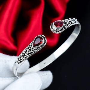 Puede incluir: Una pulsera abierta de plata pulida, con dos piedras preciosas facetadas de color rojo intenso en forma de lágrima en cada extremo. El trabajo en metal muestra intrincados patrones de volutas y pequeños detalles de cuentas.