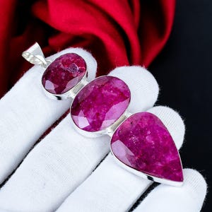 Peut inclure: Un pendentif en argent composé de trois pierres précieuses rubis facettées d'un rose profond, disposées verticalement. La pierre supérieure est ovale, la pierre centrale est un ovale plus grand et la pierre inférieure est en forme de goutte.