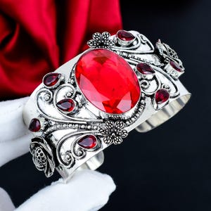 Puede incluir: Un brazalete de plata con una gran piedra roja de corte ovalado en el centro, rodeada de piedras rojas más pequeñas en forma de lágrima y detallados patrones metálicos de flores y volutas en toda la superficie de la banda ancha.