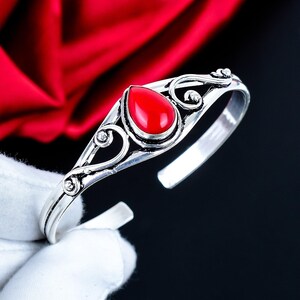 May include: A silver cuff bracelet featuring a central teardrop-shaped red stone. The metal band displays intricate swirling filigree patterns on both sides of the stone, creating a decorative openwork design along the length of the bracelet.