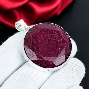 Può includere: Un grande pendente rotondo con una gemma rosso scuro sfaccettata incastonata in metallo color argento. La gemma ha una superficie lucida e riflettente. Il pendente è mostrato su sfondo nero e tessuto bianco.