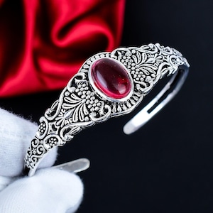 Op de afbeelding: Een zilverkleurige manchetarmband met een ovale, dieprode edelsteen in het midden. De armband heeft ingewikkelde bloemen- en wijnstokpatronen langs de band. De edelsteen is gezet in een zilveren bezel en de armband heeft een open ontwerp.