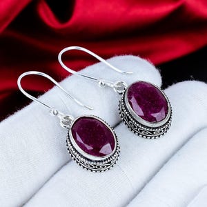 Può includere: Orecchini in argento con pietre preziose ovali rosso scuro. Le pietre sono incastonate in una cornice d'argento decorativa e decorata. Gli orecchini hanno una chiusura a gancio. Sfondo rosso.