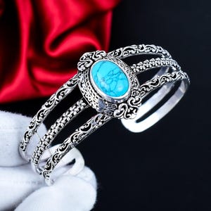 May include: Silver cuff bracelet with an oval turquoise stone. The bracelet has three bands with floral and scrollwork. The turquoise stone is a bright blue. The bracelet is approximately 15 cm in circumference.
