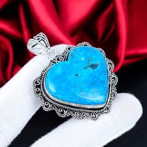 Puede incluir: Un colgante en forma de corazón con una vibrante piedra turquesa. La piedra está engastada en un marco plateado adornado con una fianza decorativa. El colgante se sostiene sobre una superficie blanca con un fondo rojo.
