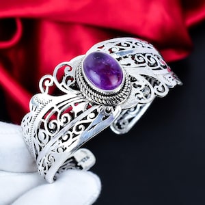 Peut inclure: Un bracelet manchette en argent avec une pierre précieuse ovale violet foncé au centre. Le bracelet présente des motifs ajourés complexes et une bordure texturée autour de la pierre précieuse. Le fond est un tissu rouge foncé.