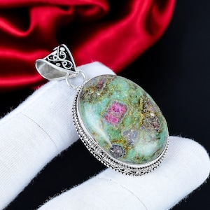 Pode incluir: Pingente oval com uma grande pedra preciosa verde e vermelha. A pedra preciosa é colocada em um metal prateado com um design ornamentado nas bordas e uma fiança decorativa. O fundo é uma mistura de vermelho e preto.