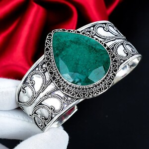 Può includere: Un bracciale a fascia in argento con una grande pietra preziosa verde sfaccettata a forma di pera al centro. La fascia metallica è decorata con intricati motivi a volute e filigrana in rilievo che coprono l'intera superficie del bracciale.