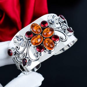 May include: A wide silver cuff bracelet featuring four central oval amber cabochons arranged in a cluster, surrounded by smaller round and pear-shaped red garnet stones, accented with intricate silver floral and swirling vine metalwork.