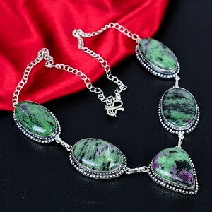 Puede incluir: Un collar de plata con piedras preciosas ovaladas y en forma de lágrima de color verde y púrpura. Las piedras están engastadas en un bisel de plata con un borde de cuentas y conectadas por una cadena de plata. El collar se exhibe sobre un fondo de tela roja.